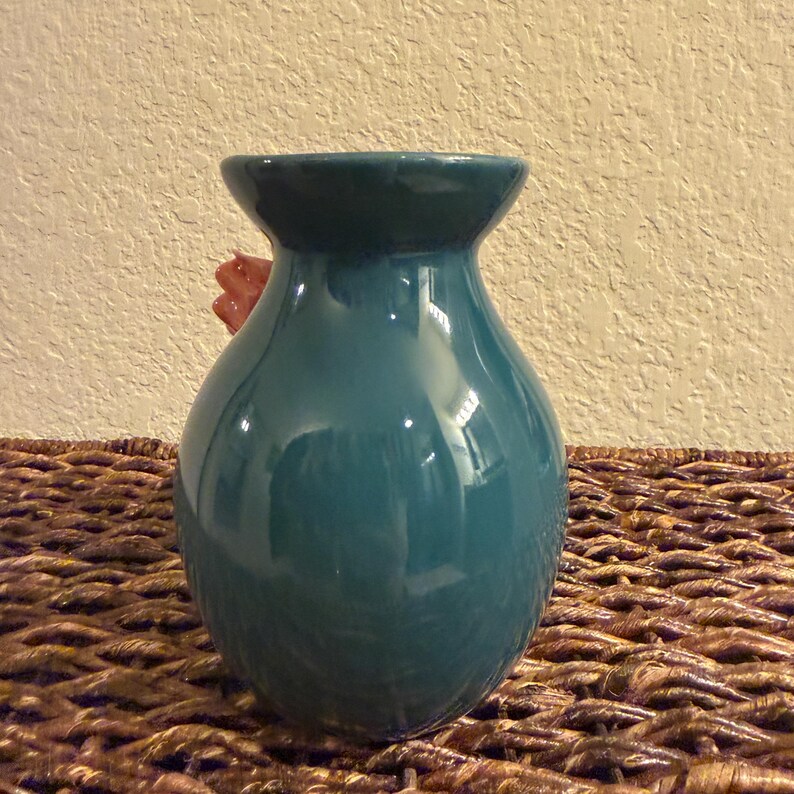 May include: A teal ceramic vase with a glossy finish. The vase has a rounded body and a flared opening. A red flower is partially visible inside the vase. The vase is sitting on a woven brown surface.