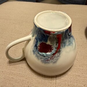 May include: A ceramic mug with a white base and a handle. The mug features a design with blue, red, and white swirls. The interior is white. The mug is sitting on a beige surface.