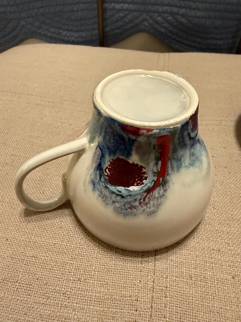May include: A ceramic mug with a white base and a handle. The mug features a design with blue, red, and white swirls. The interior is white. The mug is sitting on a beige surface.