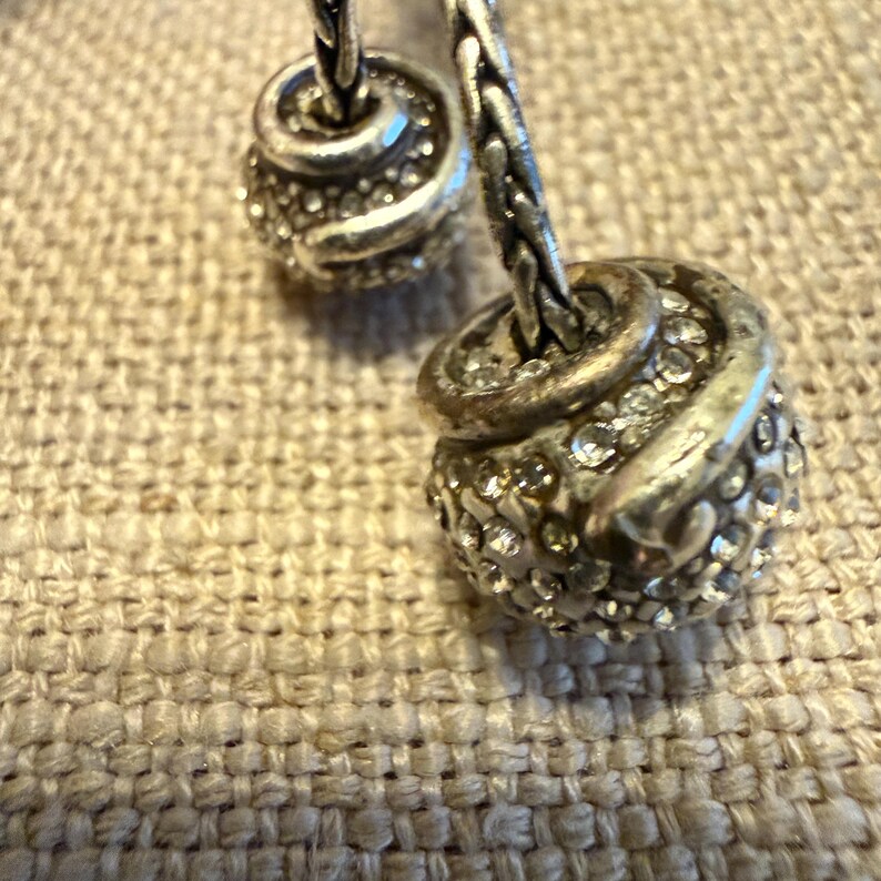 May include: A silver-toned bracelet featuring two spherical charms with a braided metal texture. Each charm is encrusted with numerous small, clear, reflective stones that catch the light, attached to a woven silver-toned chain link band.