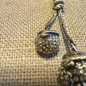 May include: A silver-toned necklace featuring two spherical pendants encrusted with clear, sparkling rhinestones. The pendants are attached to a braided silver-toned chain that connects at a central knot detail, creating a textured, metallic look.