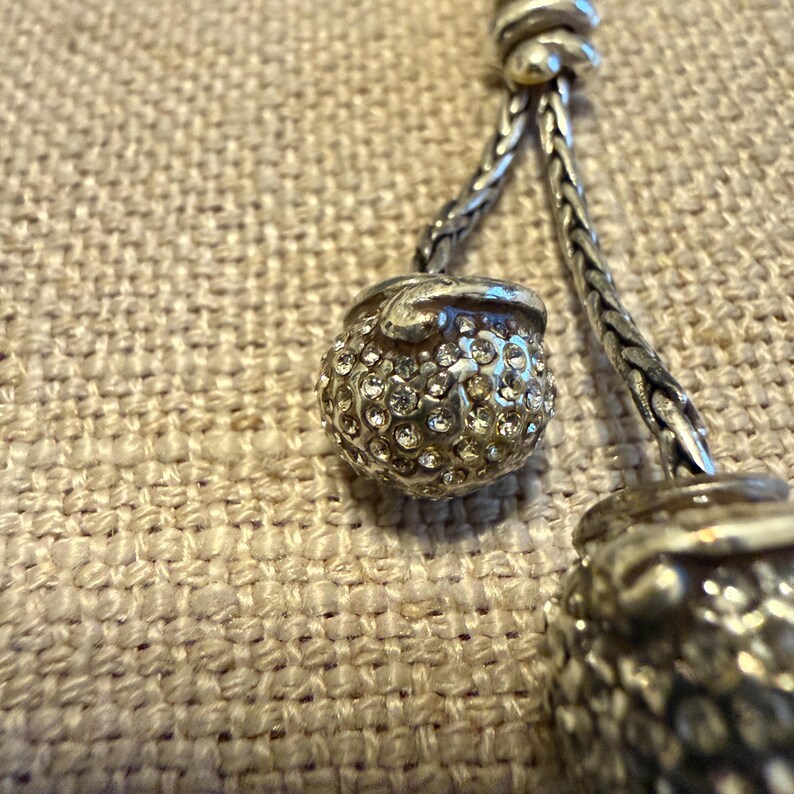 May include: A silver-toned necklace featuring two spherical pendants encrusted with clear, sparkling rhinestones. The pendants are attached to a braided silver-toned chain that connects at a central knot detail, creating a textured, metallic look.