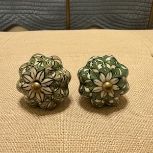 May include: Two decorative ceramic cabinet knobs. Each knob is a flower shape with a green and white floral pattern. The center of each knob is a gold-colored metal.