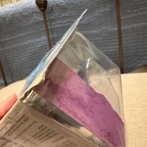 以下が含まれることがあります： 紫と白のデザインの透明なプラスチックパッケージ。パッケージには小さな玩具のフィギュアが入っています。パッケージの裏には「4歳以上」の文字があります。