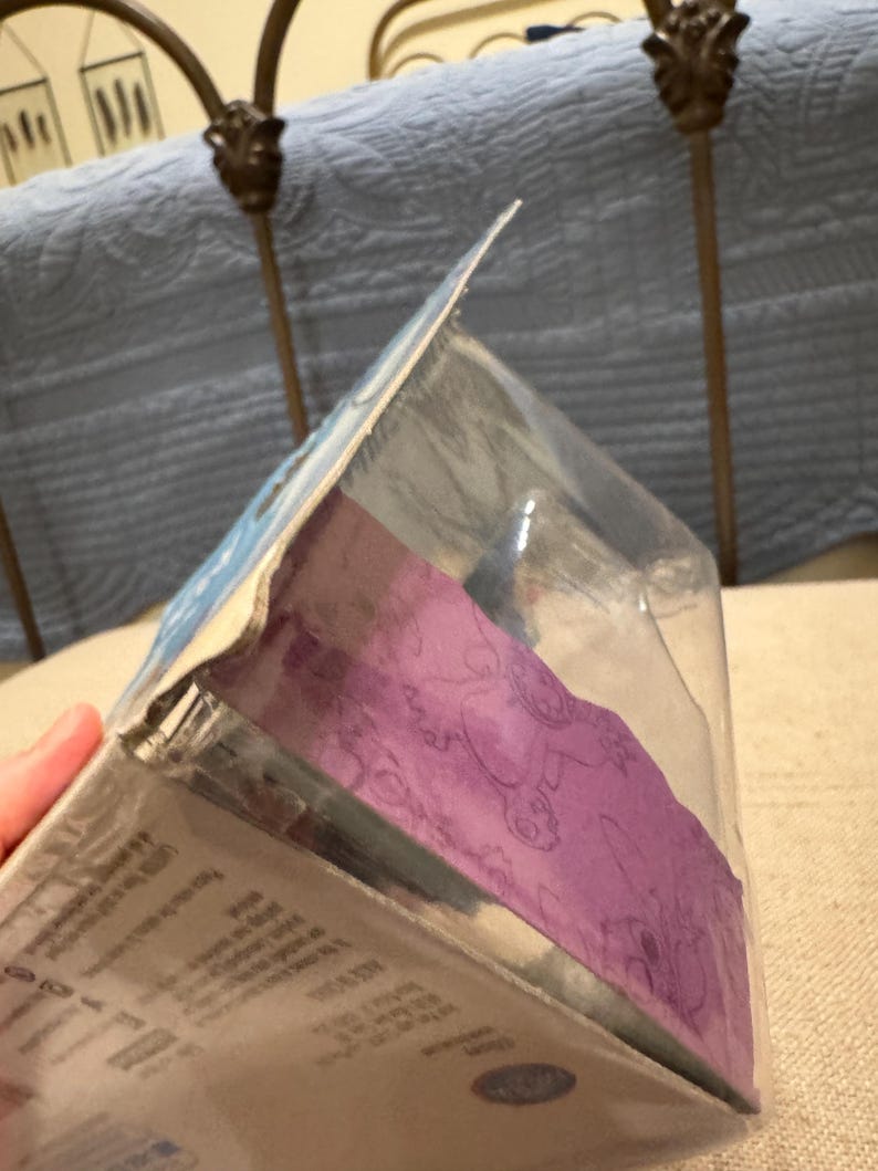 以下が含まれることがあります： 紫と白のデザインの透明なプラスチックパッケージ。パッケージには小さな玩具のフィギュアが入っています。パッケージの裏には「4歳以上」の文字があります。