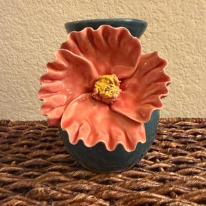 May include: A teal ceramic vase with a large, coral-colored flower design. The flower has ruffled petals and a yellow center. The vase is sitting on a woven surface.