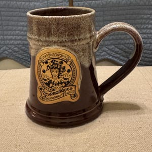 May include: A brown ceramic mug with a white glaze. The mug has a handle and a logo on the front that says "Step Back in Time 2008 Scarborough Renaissance Festival".