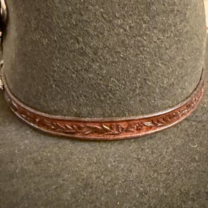 May include: Close-up of a dark green felt hat with a brown leather band. The leather band is adorned with an embossed floral pattern. The hat's brim is slightly curved, and the overall design suggests a classic, rustic style.