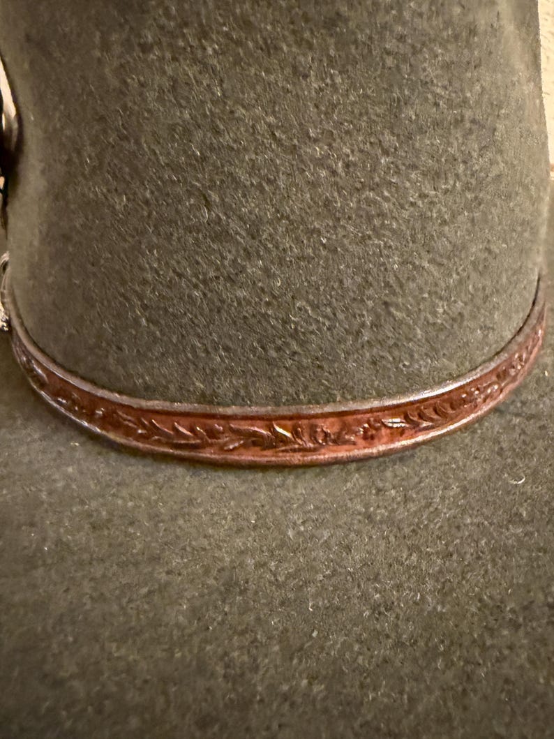 May include: Close-up of a dark green felt hat with a brown leather band. The leather band is adorned with an embossed floral pattern. The hat's brim is slightly curved, and the overall design suggests a classic, rustic style.