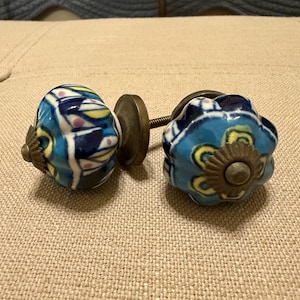 May include: Two decorative ceramic cabinet knobs. One knob is blue, yellow, and white with a floral design. The other knob is blue with yellow floral accents. Both knobs have a brass-colored base and screw.
