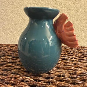 May include: A teal ceramic vase with a glossy finish and a coral-colored, wing-shaped accent. The vase has a rounded body and a flared rim. The wing detail adds a unique, decorative touch to the vessel.