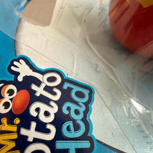 Może przedstawiać: Zbliżenie opakowania zabawki Mr. Potato Head. Opakowanie jest niebiesko-białe ze słowami "Mr. Potato Head" w kolorze białym i niebieskim. Czerwona i ż&oacute;łta zabawka jest widoczna w opakowaniu.