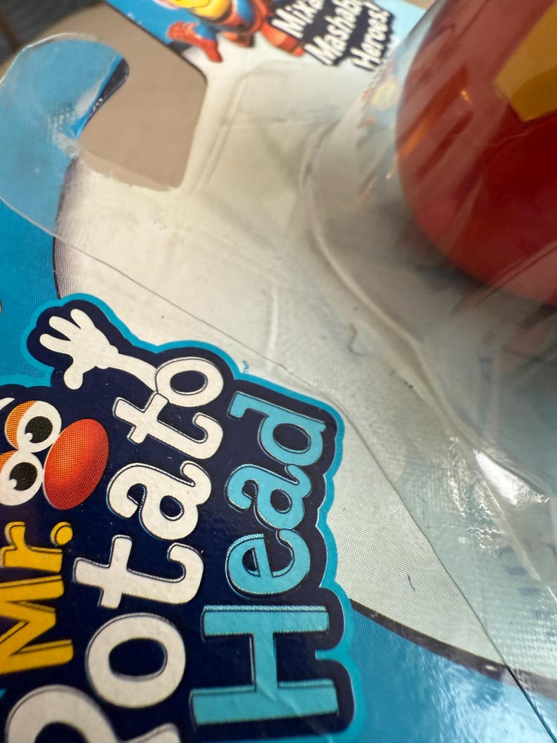 Może przedstawiać: Zbliżenie opakowania zabawki Mr. Potato Head. Opakowanie jest niebiesko-białe ze słowami "Mr. Potato Head" w kolorze białym i niebieskim. Czerwona i ż&oacute;łta zabawka jest widoczna w opakowaniu.
