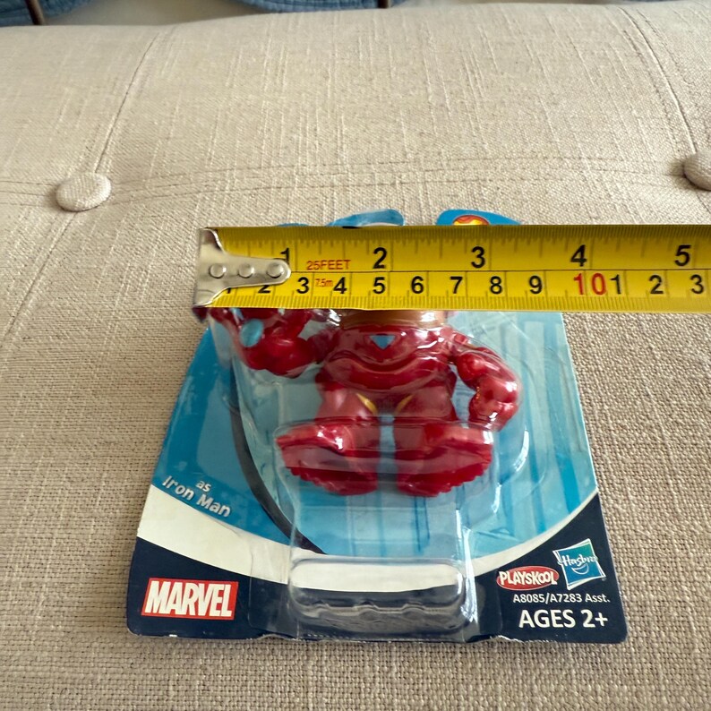 Może przedstawiać: Czerwona figurka Iron Mana, o wysokości około 10 cm, w oryginalnym opakowaniu. Opakowanie zawiera logo Marvel i markę Playskool. Figurka przeznaczona jest dla dzieci w wieku 2+.