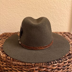 May include: A dark olive green fedora hat with a wide brim. The hat features a brown leather band with embossed detailing and a decorative feather accent. The hat is displayed on a woven basket.