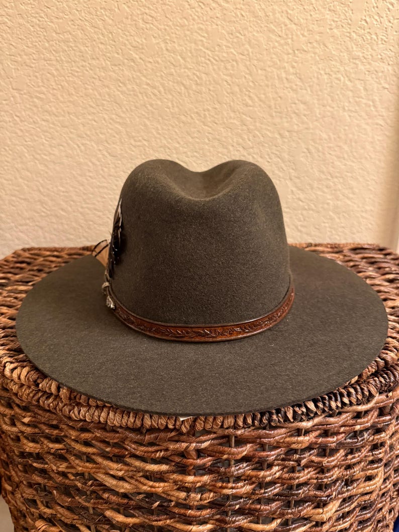 May include: A dark olive green fedora hat with a wide brim. The hat features a brown leather band with embossed detailing and a decorative feather accent. The hat is displayed on a woven basket.