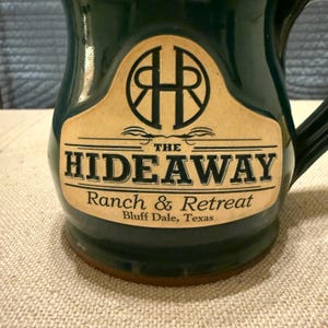 May include: A dark green ceramic mug with a handle. The mug features a tan label with the words "THE HIDEAWAY Ranch & Retreat Bluff Dale, Texas" in black text. The label also has a logo with the letters "H" and "R".