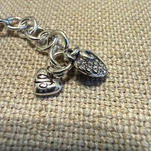 May include: A silver-toned metal chain bracelet featuring two heart-shaped charms attached to a circular link. One charm is smooth with an engraved script letter, while the other heart charm is textured and encrusted with small clear crystals.