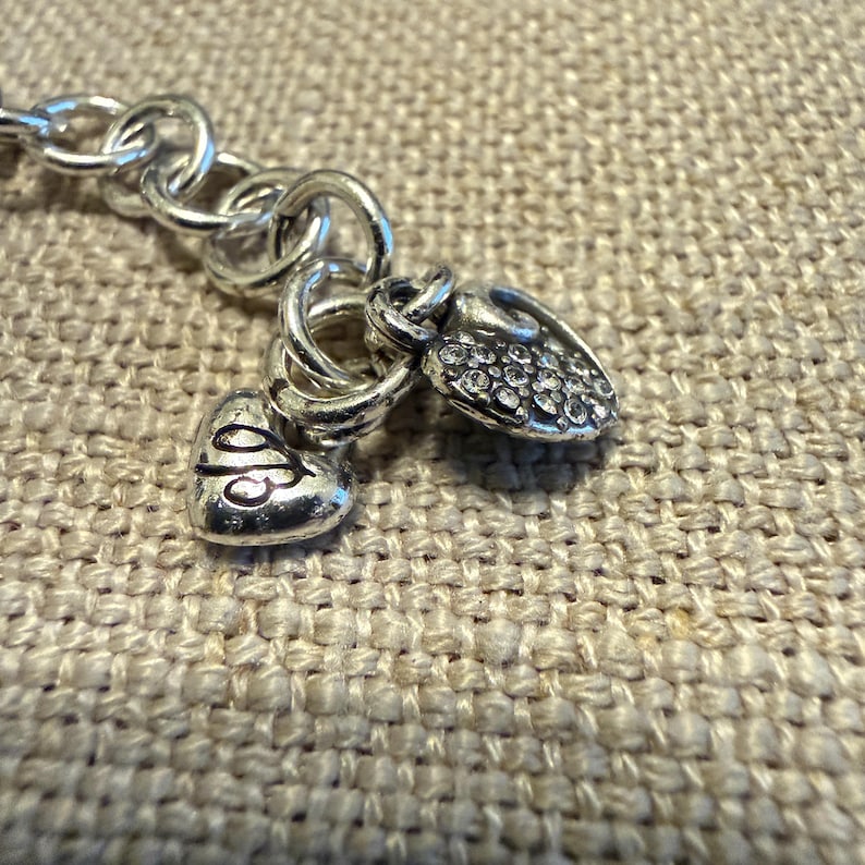 May include: A silver-toned metal chain bracelet featuring two heart-shaped charms attached to a circular link. One charm is smooth with an engraved script letter, while the other heart charm is textured and encrusted with small clear crystals.