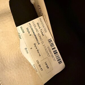 May include: A tag with the text "OVERLAND NOMAD OL 3 P.NCH FLT" and "Large" size. The tag is attached to a light-colored fabric. The color is "OLIVE". The style number is PF21500NOM300LLG.