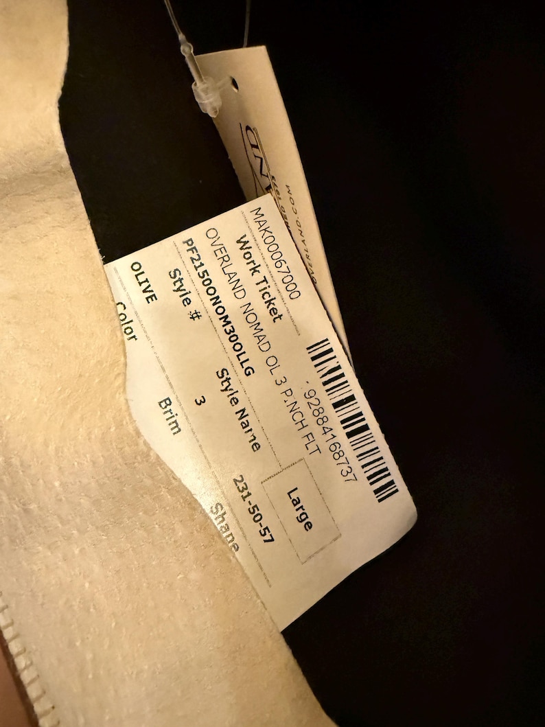 May include: A tag with the text "OVERLAND NOMAD OL 3 P.NCH FLT" and "Large" size. The tag is attached to a light-colored fabric. The color is "OLIVE". The style number is PF21500NOM300LLG.