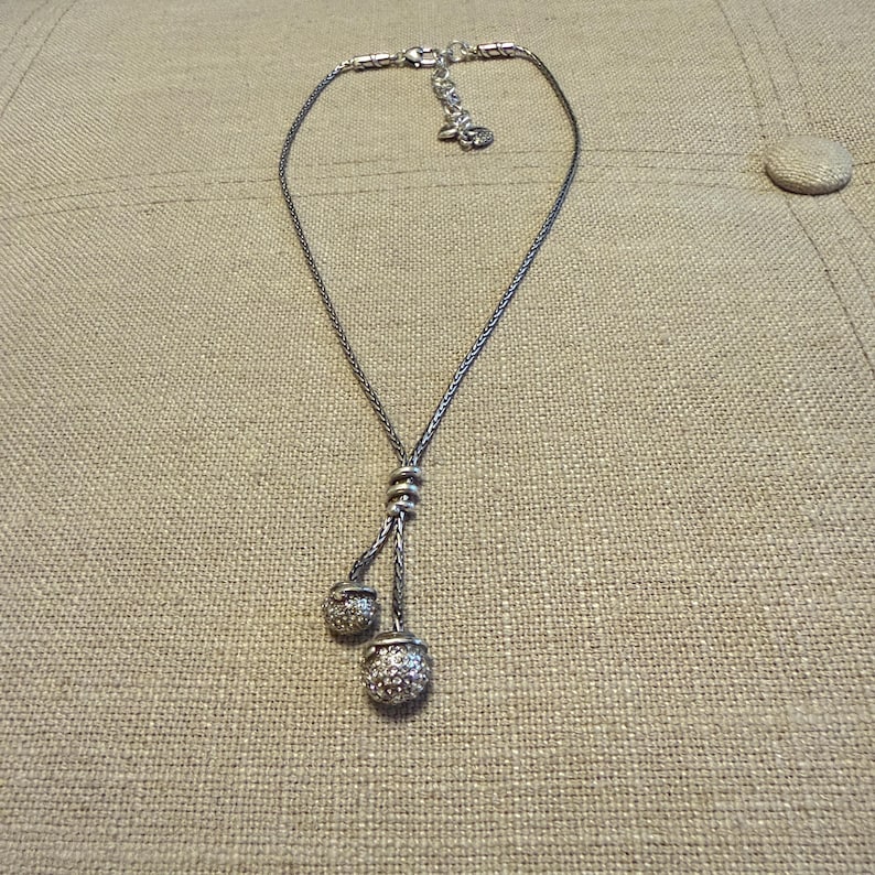 May include: A silver-toned lariat necklace featuring a delicate woven chain and two dangling spherical charms. The charms are encrusted with small, clear, reflective stones. The necklace includes a lobster claw clasp and an adjustable extender chain.