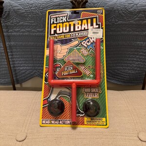 May include: A new Championship Flick Football game for 1-2 players. The game includes a red goal post, two black footballs, and a game board with the text "Head2Head Action!" and "Two Skill Levels!"