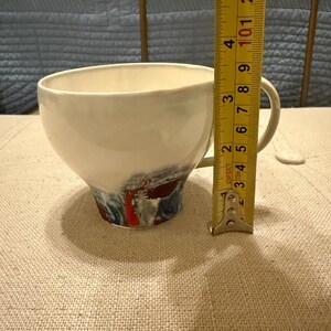 May include: A white ceramic mug with a black handle and the text "But First, Coffee" in a bold, black font. The mug is set against a blurred background, suggesting a cozy setting.
