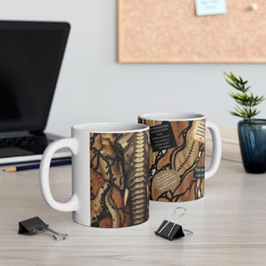 May include: White ceramic coffee mug featuring a detailed illustration of an elephant's face and trunk, overlaid with a snakeskin pattern in shades of brown and black. The mug also has text on it.