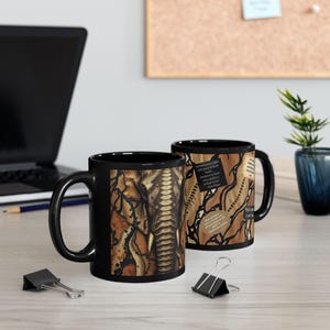 May include: Two black coffee mugs with a unique design. One mug features a close-up of an elephant's face with a snakeskin pattern. The other mug has a snakeskin pattern with text. The mugs are on a light-colored wooden surface.