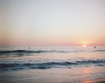 Könnte beinhalten: Eine malerische Aussicht auf einen Strand bei Sonnenuntergang mit ein paar Personen im Wasser. Der Himmel ist in sanftem Rosa und Orange gehalten, und das Wasser ist ruhig blau.