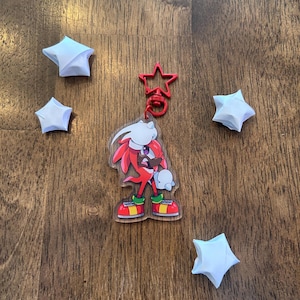 May include: A red and white cartoon character keychain with a red star charm. The character is standing with its arms crossed and has a white stripe on its head. The keychain is made of clear acrylic and is surrounded by white origami stars.