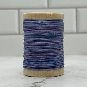 May include: A wooden spool wrapped with thin, multi-colored thread featuring alternating horizontal bands of muted blue and soft purple tones. The thread is wound tightly around the cylindrical wooden core, creating a textured, layered appearance.
