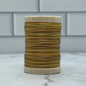 May include: A wooden spool of thread, tightly wound with threads in shades of yellow, gold, and brown. The thread is layered, creating a textured effect. The spool is set against a white background.