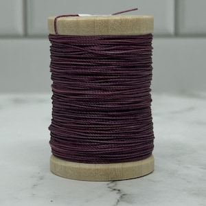 May include: A wooden spool tightly wound with dark purple thread. The thread is a deep, rich shade of purple, possibly waxed linen. The spool is made of light-colored wood. The thread is suitable for sewing or crafting projects. The thread is tightly wound.