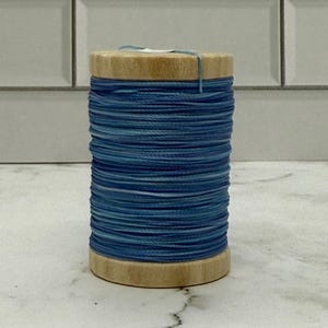 May include: A wooden spool of blue thread. The thread is tightly wound around the spool, with varying shades of blue creating a textured appearance. A small length of thread extends from the top. The spool is sitting on a marble surface.