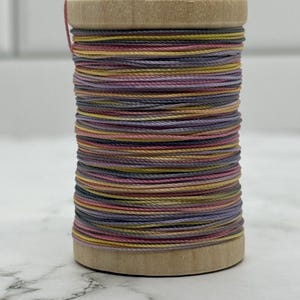 May include: A wooden spool wrapped with multi-coloured thread in shades of dusty pink, muted yellow, lavender, and slate grey. The thread is wound tightly around the cylindrical wooden core, creating a layered, striped appearance of soft pastel tones.