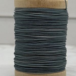 May include: A wooden spool wrapped with a continuous length of slate blue thread. The thread is wound tightly around the cylindrical wooden core, creating a uniform texture. A single loose end of the blue thread extends from the top of the spool.
