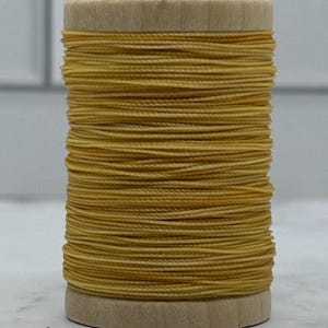 May include: A cylindrical wooden spool wrapped with a continuous length of mustard yellow thread. The thread is wound tightly around the centre of the spool, creating a uniform texture with visible horizontal lines across the entire surface.