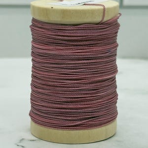 May include: A wooden spool wrapped with a continuous length of dusty rose coloured thread. The thread is wound tightly around the cylindrical wooden core, creating a uniform texture. A small white label is partially visible on the top surface.