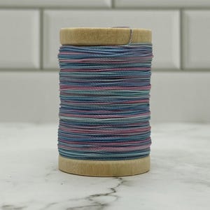 May include: A wooden spool wrapped with thin, multi-colored thread featuring alternating horizontal bands of dusty blue, soft pink, and muted teal. The spool has a natural light wood finish with flat circular ends at the top and bottom.