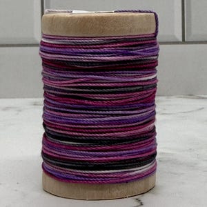 Könnte beinhalten: Eine Holzspule, eng gewickelt mit farbigem Garn in Lila-, Rosa-, Grau- und Weißtönen. Der Faden ist sauber geschichtet und erzeugt ein ansprechendes Muster. Ideal für Bastel- und Nähprojekte.