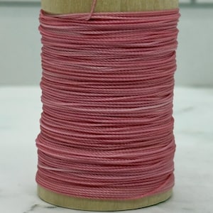 May include: A cylindrical wooden spool wrapped with a continuous length of dusty pink thread. The spool features a small white label on the top edge with partially visible black text, and the thread is wound tightly in horizontal layers.