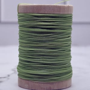 May include: A wooden spool wrapped with sage green thread. The thread is wound tightly around the cylindrical wooden core, creating a uniform texture. The spool has flat circular ends and a smooth, light-coloured wood grain finish throughout.
