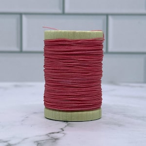 May include: A spool of red thread, tightly wound, sits on a light green base. The thread is a vibrant shade of red, and the spool is set against a white and grey background. This image is ideal for craft and sewing projects.