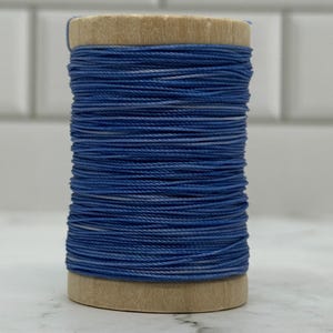 May include: A wooden spool wound with blue thread. The thread is a mix of solid and variegated shades of blue, creating a textured appearance. The spool is made of light-coloured wood.