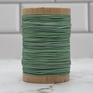 May include: A wooden spool wrapped with sage green thread. The thread is wound tightly around the cylindrical wooden core, creating a uniform texture. The spool features natural light-coloured wood grain at the top and bottom ends of the cylinder.