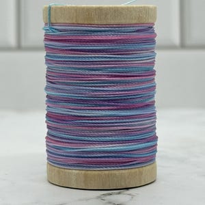 May include: A wooden spool wrapped with thin cord in alternating horizontal bands of light blue, lavender, and magenta. The spool features a natural light-colored wood finish on the top and bottom ends, with the colorful thread wound tightly around the center.