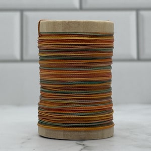 May include: A wooden spool wrapped with multiple strands of thread in alternating shades of orange, yellow, brown, and muted green. The thread is wound horizontally around the cylindrical wooden core, creating a layered, multi-coloured appearance.