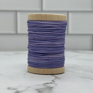 May include: A spool of purple thread with a wooden base and top. The thread is tightly wound, creating a cylindrical shape. The color of the thread varies slightly, with hints of lavender and white. Ideal for sewing, embroidery, or other crafts.